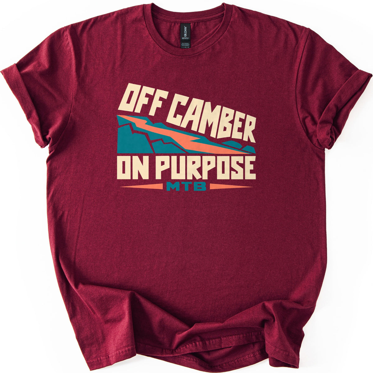 Off Camber On Purpose Comfort Colors T-shirt in Maroon, laid flat. Cream block letters read Off Camber On Purpose around a teal mountain slope cut by a diagonal orange zigzag trail, with MTB in teal over a thin orange line. The sleeves are cuffed on a white background.
