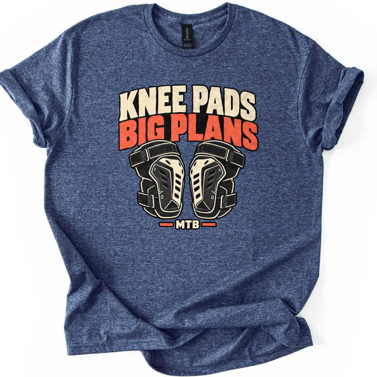 Knee Pads Big Plans Comfort Colors T-shirt in Heather Navy, laid flat. The words Knee Pads Big Plans in cream and orange-red block letters arch above a pair of black and cream knee pads, with MTB between orange bars below. The shirt has rolled sleeves on a white background.