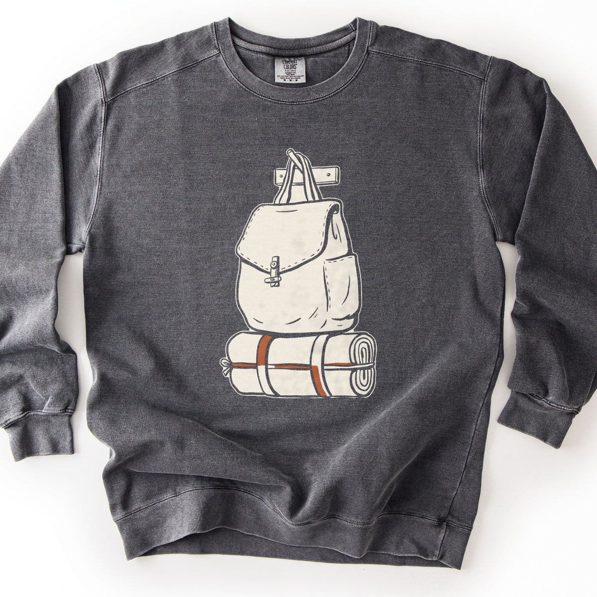 Backpack And Bedroll Comfort Colors crewneck sweatshirt in Pepper, laid flat. A cream canvas backpack with a toggle clasp hangs by its straps from a wall peg above a rolled bedroll tied with rust-orange straps. No words are printed. Shown on a white background.