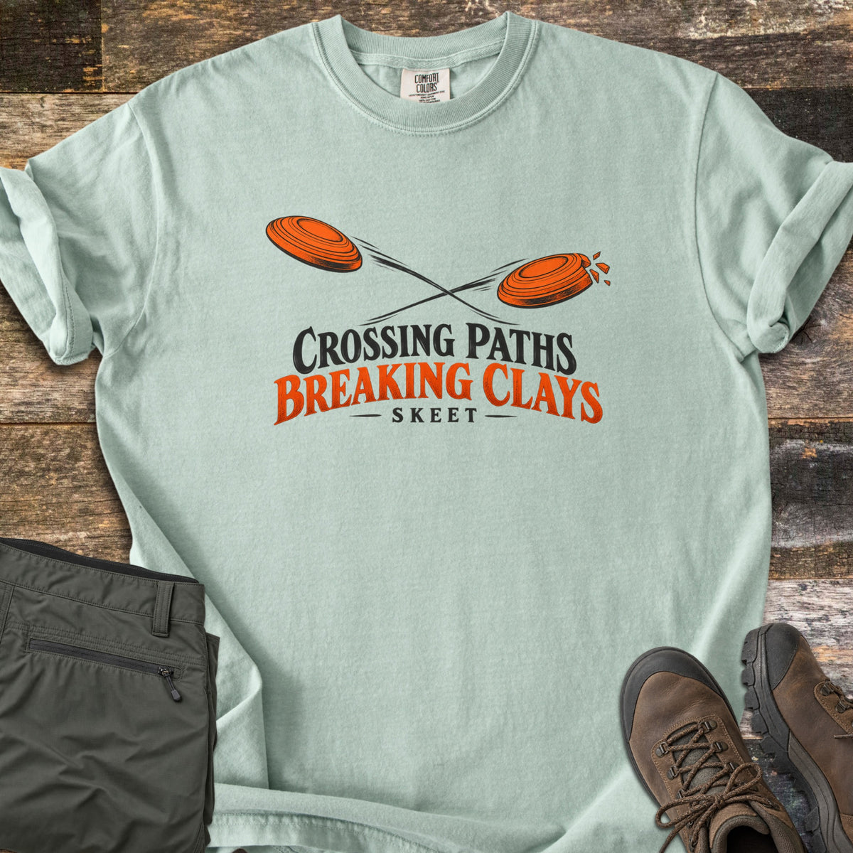 Crossing Paths Breaking Clays Comfort Colors T-shirt in Bay, laid flat. Two orange clay targets cross paths, one shattering, above the words Crossing Paths Breaking Clays and Skeet. Sleeves rolled, on dark barn wood with olive pants and brown hiking boots.