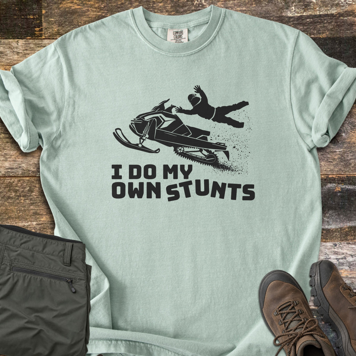 I Do My Own Stunts Comfort Colors T-shirt in Bay, laid flat. A black silhouette of a rider thrown from a snowmobile mid-jump, with snow spraying from the track, above the words I Do My Own Stunts. Olive cargo pants and brown hiking boots lie on rustic wood planks.