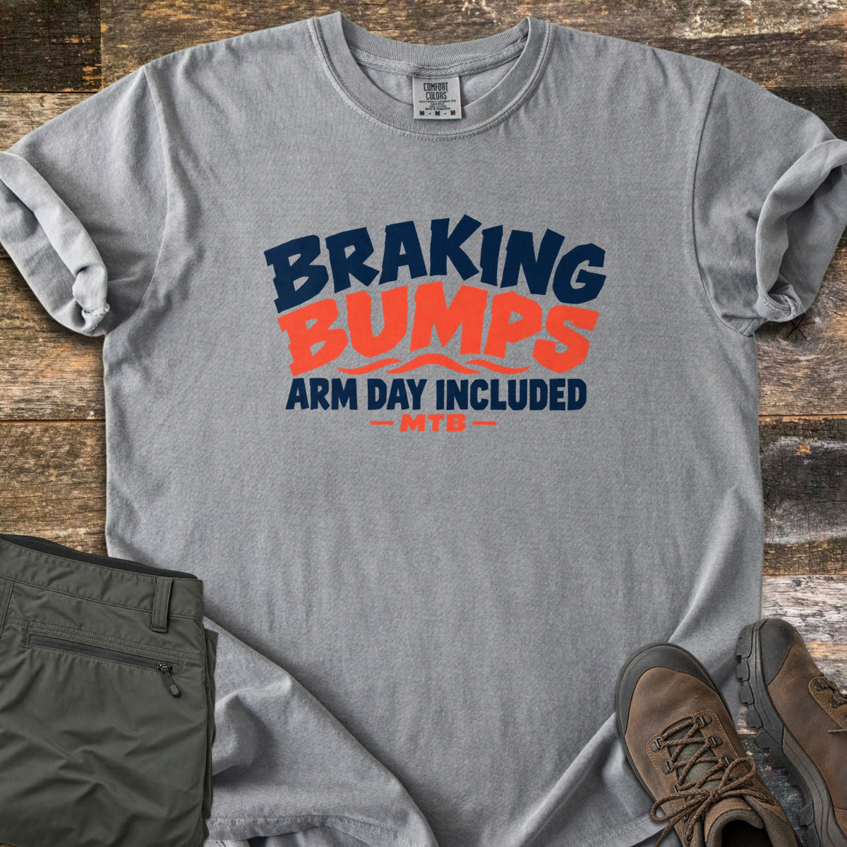 Braking Bumps Arm Day Included Comfort Colors T-shirt in Grey, laid flat. Bold navy and orange text reads Braking Bumps, Arm Day Included, MTB, with wavy orange lines under Bumps. It rests on a wood floor beside olive cargo pants and brown hiking boots.