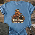 Spa Day Trail Edition Comfort Colors T-shirt in Blue Jean, laid flat. A smiling cartoon bear in a camo helmet rides a muddy orange ATV through splashing mud above the words Spa Day Trail Edition. Shown on a rustic wood floor with olive cargo pants and brown hiking boots.