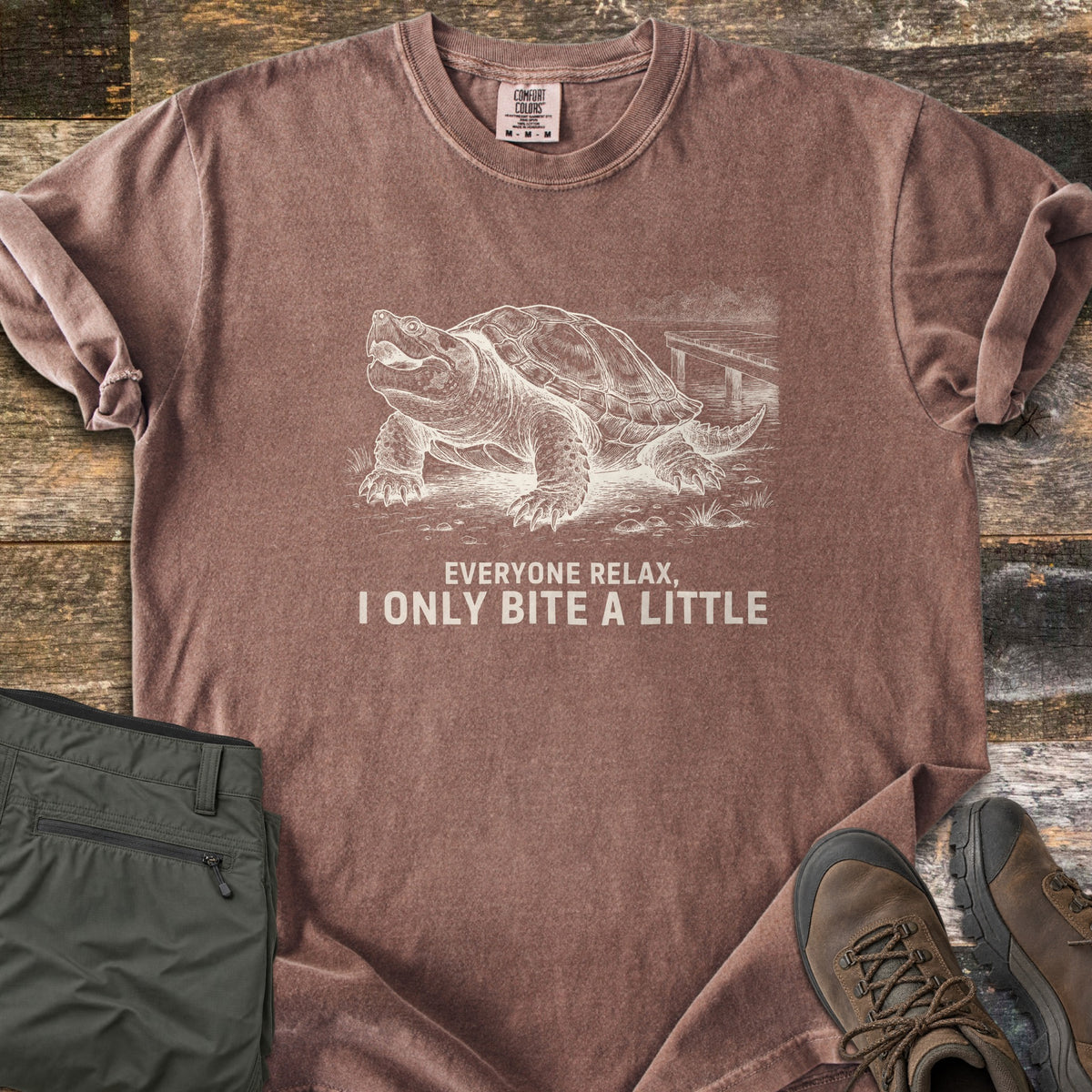 Everyone Relax I Only Bite A Little Comfort Colors T-shirt in Espresso, laid flat. A cream sketch of a snapping turtle with open mouth on a lakeshore by a wooden dock, above the words Everyone relax, I only bite a little. Olive hiking pants and brown hiking boots sit on a weathered wood floor.