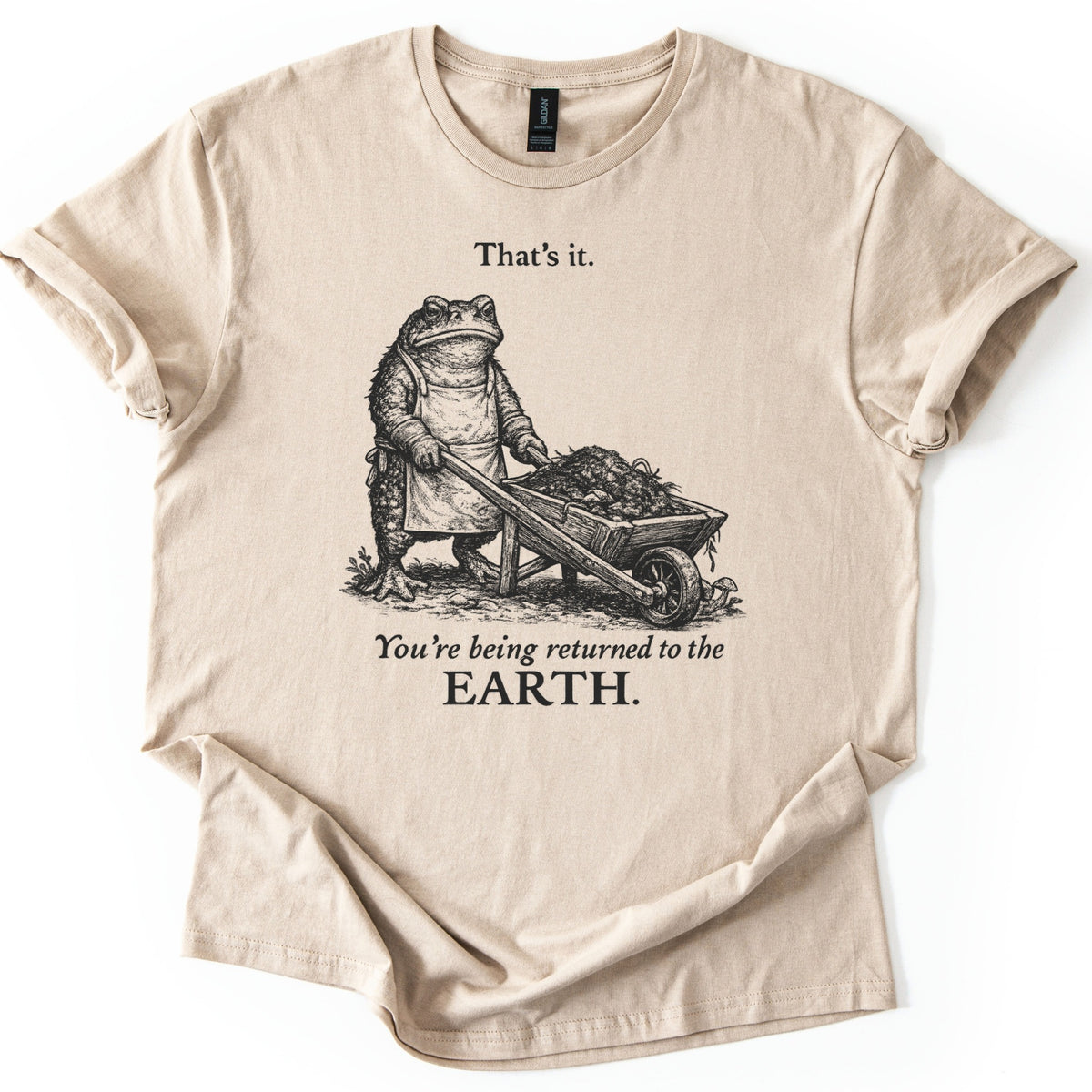 Toad Compost Comfort Colors T-shirt in Sand, laid flat. A black ink etching of a frowning toad in an apron pushing a wheelbarrow of compost, between the words That's it. and You're being returned to the EARTH. Sleeves are cuffed on a white background.