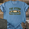 The Stern Takes the Blame Comfort Colors T-shirt in Blue Jean, laid flat. A top-down green canoe with a red arrow marking the back seat as ME sits between the words The Stern and Takes the Blame, with two wooden paddles below. Olive hiking pants and brown boots lie on weathered wood.