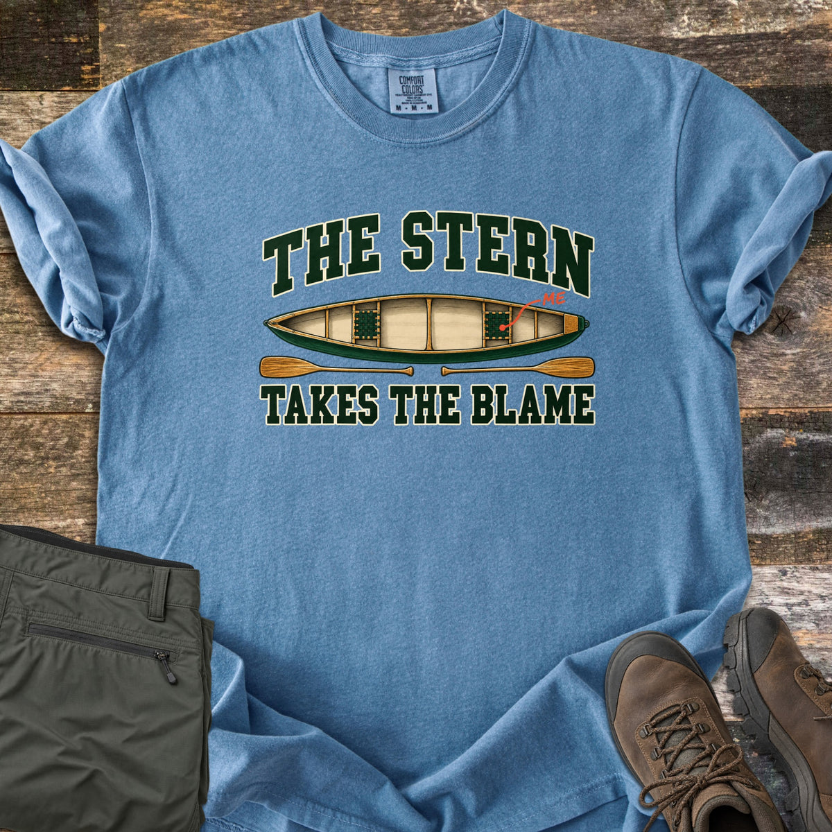The Stern Takes the Blame Comfort Colors T-shirt in Blue Jean, laid flat. A top-down green canoe with a red arrow marking the back seat as ME sits between the words The Stern and Takes the Blame, with two wooden paddles below. Olive hiking pants and brown boots lie on weathered wood.