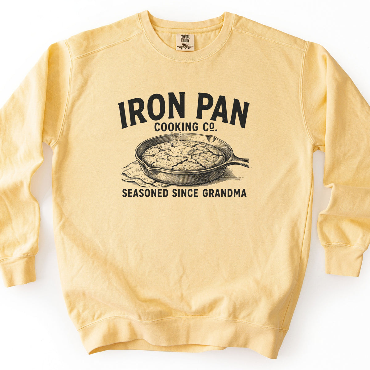 Iron Pan Cooking Co Comfort Colors crewneck sweatshirt in Butter, laid flat. Black print of a cast iron skillet holding cornbread topped with melting butter, resting on a cloth, with the words Iron Pan Cooking Co. above and Seasoned Since Grandma below. Shown on a white background.