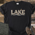 Lake Comfort Colors T-shirt in Black, laid flat. The word LAKE spelled in weathered driftwood planks with a kingfisher perched on the K, above a wavy water line. Sleeves rolled, on rustic wood boards beside green hiking pants and brown hiking boots.