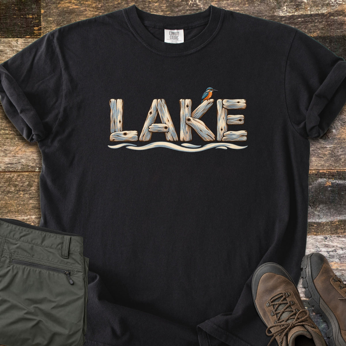 Lake Comfort Colors T-shirt in Black, laid flat. The word LAKE spelled in weathered driftwood planks with a kingfisher perched on the K, above a wavy water line. Sleeves rolled, on rustic wood boards beside green hiking pants and brown hiking boots.