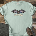 After Daylight Comfort Colors T-shirt in Bay, laid flat. A bat spreads its wings, each framing a cave view of stalactites and pine forest at dusk, above the words After Daylight. Olive hiking pants and brown hiking boots lie beside it on dark wood planks.