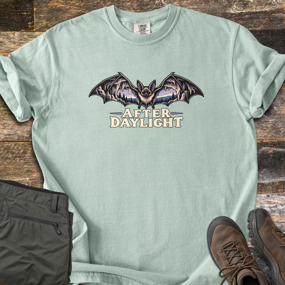 After Daylight Comfort Colors T-shirt in Bay, laid flat. A bat spreads its wings, each framing a cave view of stalactites and pine forest at dusk, above the words After Daylight. Olive hiking pants and brown hiking boots lie beside it on dark wood planks.