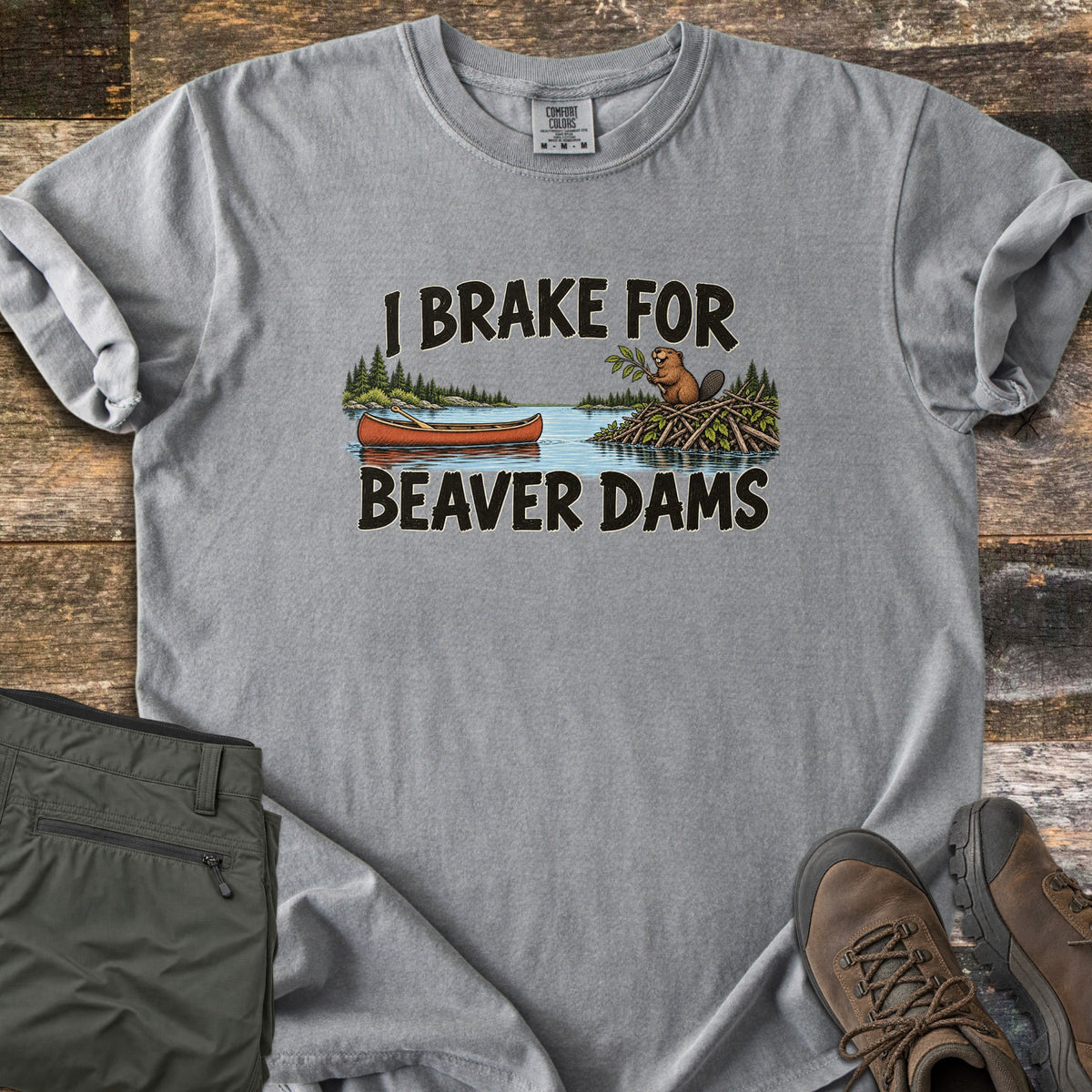 Brake For Beaver Dams Comfort Colors T-shirt in Grey, laid flat. The words I Brake For Beaver Dams frame a lake scene with a red canoe and a beaver on its stick dam among pine trees. Sleeves rolled, on weathered wood with folded green hiking pants and brown hiking boots.