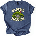 Older & Mossier Comfort Colors T-shirt in Heather Navy, laid flat. A smiling gray rock draped in green moss wears a red party hat with white dots, between the words Older & and Mossier in cream bubble letters. The shirt lies on a white background with its sleeves cuffed.