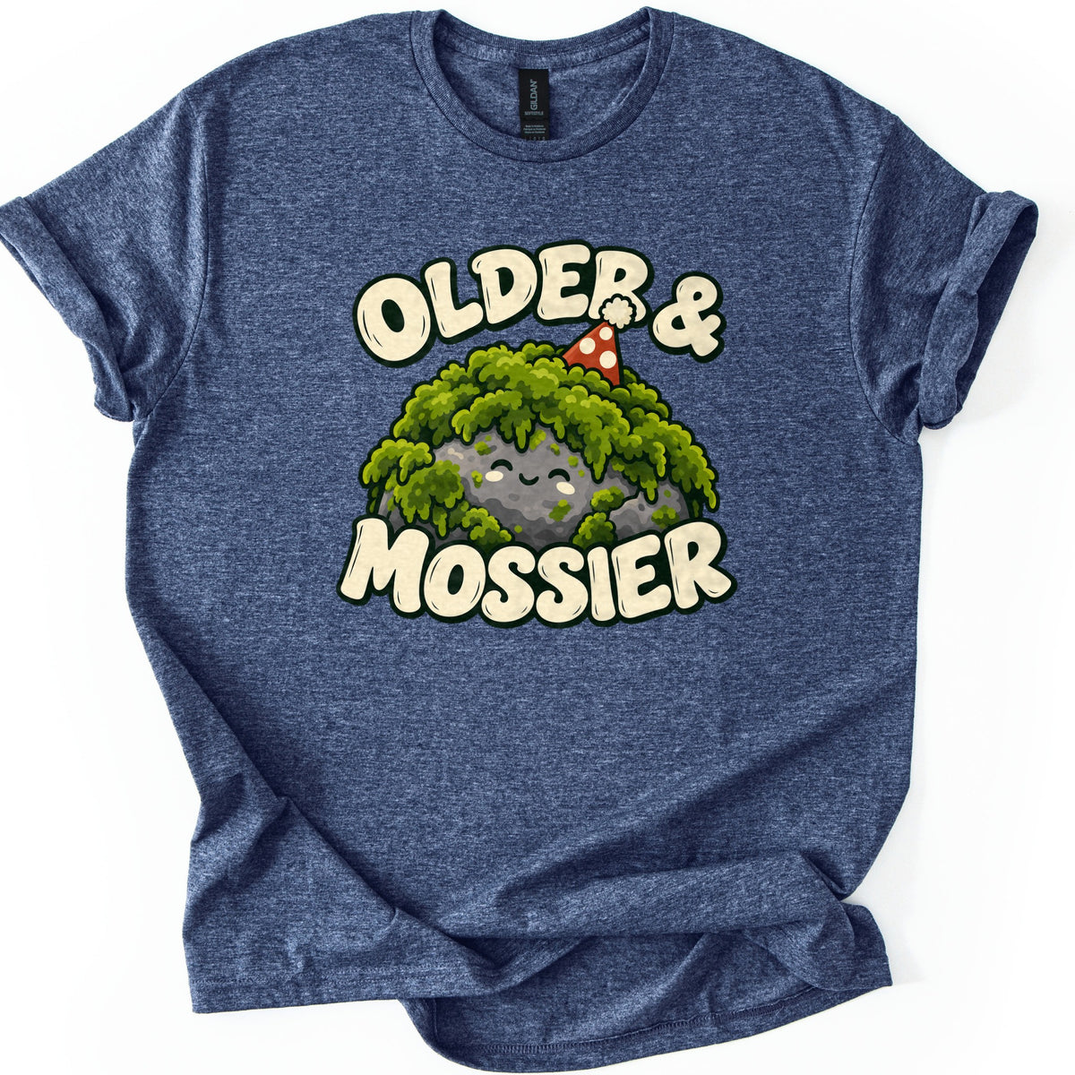 Older & Mossier Comfort Colors T-shirt in Heather Navy, laid flat. A smiling gray rock draped in green moss wears a red party hat with white dots, between the words Older & and Mossier in cream bubble letters. The shirt lies on a white background with its sleeves cuffed.