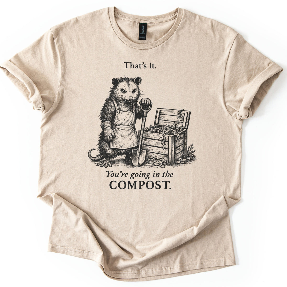 Going In The Compost Possum Comfort Colors T-shirt in Sand, laid flat. A scowling possum in an apron and work glove leans on a shovel beside an open wooden compost bin, between the words That's it. and You're going in the COMPOST. Black print, shirt on white with cuffed sleeves.