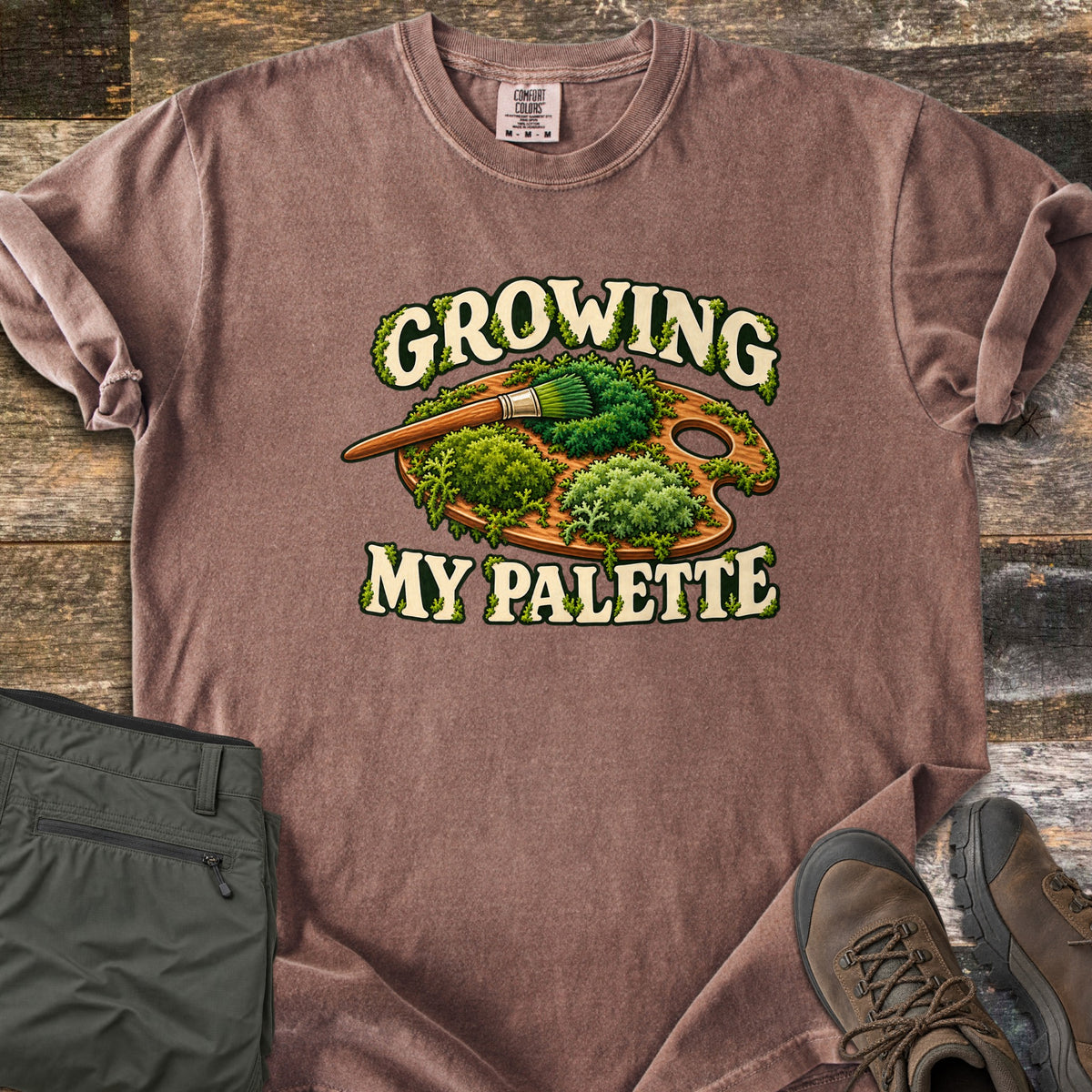 Growing My Palette Comfort Colors T-shirt in Espresso, laid flat. A wooden painter's palette covered in green moss, with a brush whose bristles are moss, between the words Growing My Palette in cream letters. Olive pants and brown hiking boots lie beside it on weathered wood.