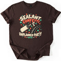 Sealant Confetti Unplanned Party Comfort Colors T-shirt in Dark Chocolate, laid flat. A cream squeeze bottle squirts sealant at a knobby bike tire amid red and green confetti dots, with the words Sealant Confetti, Unplanned Party, MTB. Sleeves are rolled, on a white background.