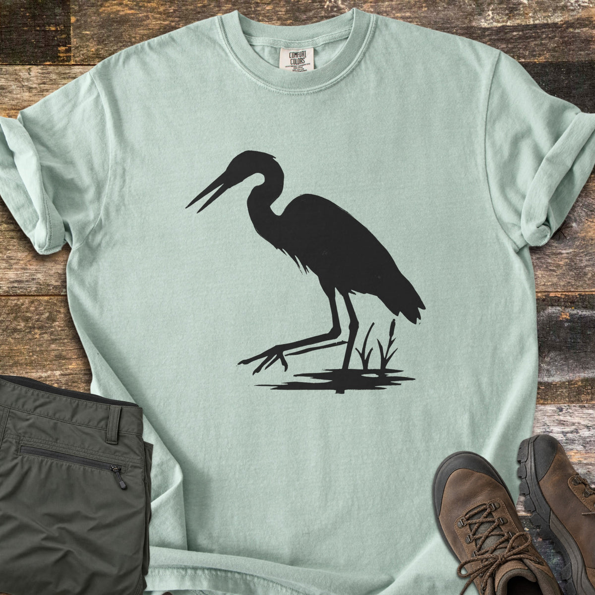Great Blue Heron Comfort Colors T-shirt in Bay, laid flat. A black silhouette of a heron wading with its beak open, one foot raised, beside cattails over a patch of water. Sleeves rolled, on weathered wood with olive hiking pants and brown hiking boots.
