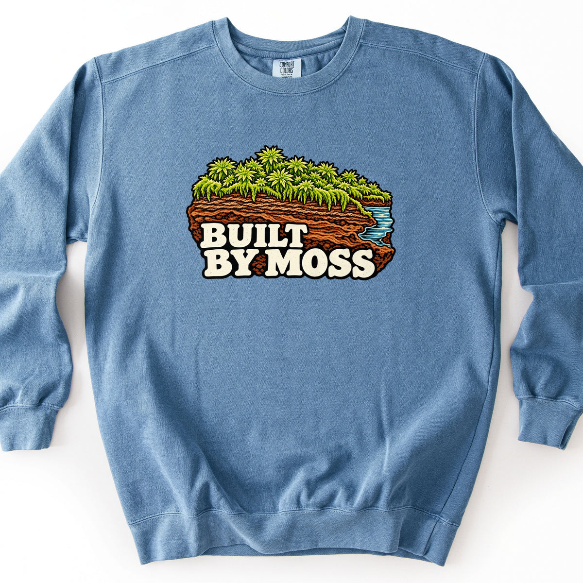 Built by Moss Comfort Colors crewneck sweatshirt in Blue Jean, laid flat. A slab of layered red-brown soil topped with bright green moss, with a small blue stream at one edge, and cream letters reading BUILT BY MOSS. It lies on a white background.