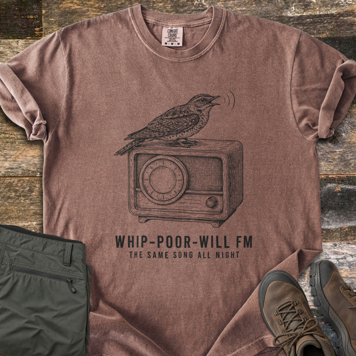Whip-Poor-Will FM Comfort Colors T-shirt in Espresso, laid flat. A sketched whip-poor-will sings atop a vintage radio above the words Whip-Poor-Will FM, The Same Song All Night. It lies with rolled sleeves on weathered wood beside folded olive pants and brown hiking boots.