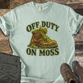 Off Duty on Moss Comfort Colors T-shirt in Bay, laid flat. A worn brown hiking boot overgrown with green moss sits between the words Off Duty and On Moss in dark green letters. Laid on weathered wood with folded olive hiking pants and brown hiking boots.