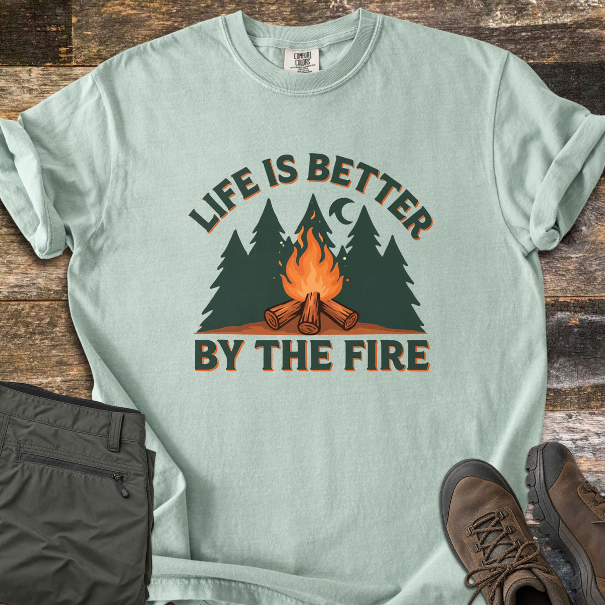 Life Is Better By The Fire Comfort Colors T-shirt in Bay, laid flat. Arched dark green text reads Life Is Better over pine trees, a crescent moon and a campfire on crossed logs, with By The Fire below. Folded olive pants and brown hiking boots sit on weathered wood.