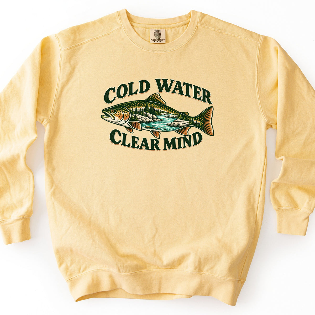 Cold Water Clear Mind Comfort Colors crewneck sweatshirt in Butter, laid flat. A spotted trout with a pine forest, rocks and a turquoise river inside its body, between the dark green words Cold Water arched above and Clear Mind below. Shown on a white background.