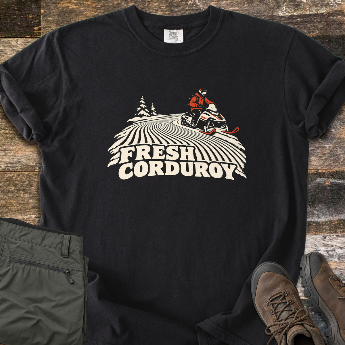 Fresh Corduroy Comfort Colors T-shirt in Black, laid flat. A person on a red and white snowmobile rides over groomed corduroy snow lines past pine trees, above the words Fresh Corduroy. It lies on weathered wood beside green pants and brown hiking boots.