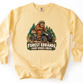 Forest Errands Four Wheel Drive Comfort Colors crewneck sweatshirt in Butter, laid flat. A grinning cartoon Bigfoot in a green helmet rides a green ATV through pine trees, a sack of pinecones on the back, above the words Forest Errands Four Wheel Drive on a log sign. Shown on a white background.