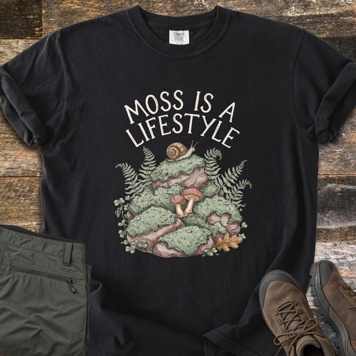 Moss Is A Lifestyle Comfort Colors T-shirt in Black, laid flat. Cream text reads Moss Is A Lifestyle above a mossy rock mound with a snail, mushrooms, ferns and clover. It lies on weathered wood beside folded olive hiking pants and brown hiking boots.