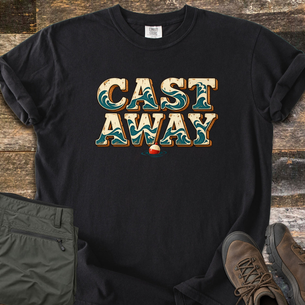 Cast Away Comfort Colors T-shirt in Black, laid flat. The words Cast Away in cream letters filled with teal waves, above a red and white fishing bobber in water. Olive cargo pants and brown hiking boots sit beside it on weathered wood planks.