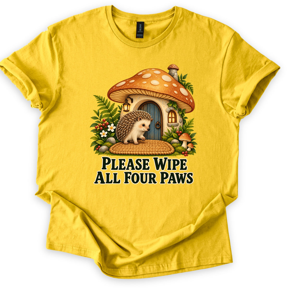 Please Wipe All Four Paws Comfort Colors T-shirt in Daisy, laid flat. A hedgehog stands on a woven doormat outside a mushroom cottage with an orange spotted cap and blue door, framed by ferns and berries. Below, dark green text reads Please Wipe All Four Paws.