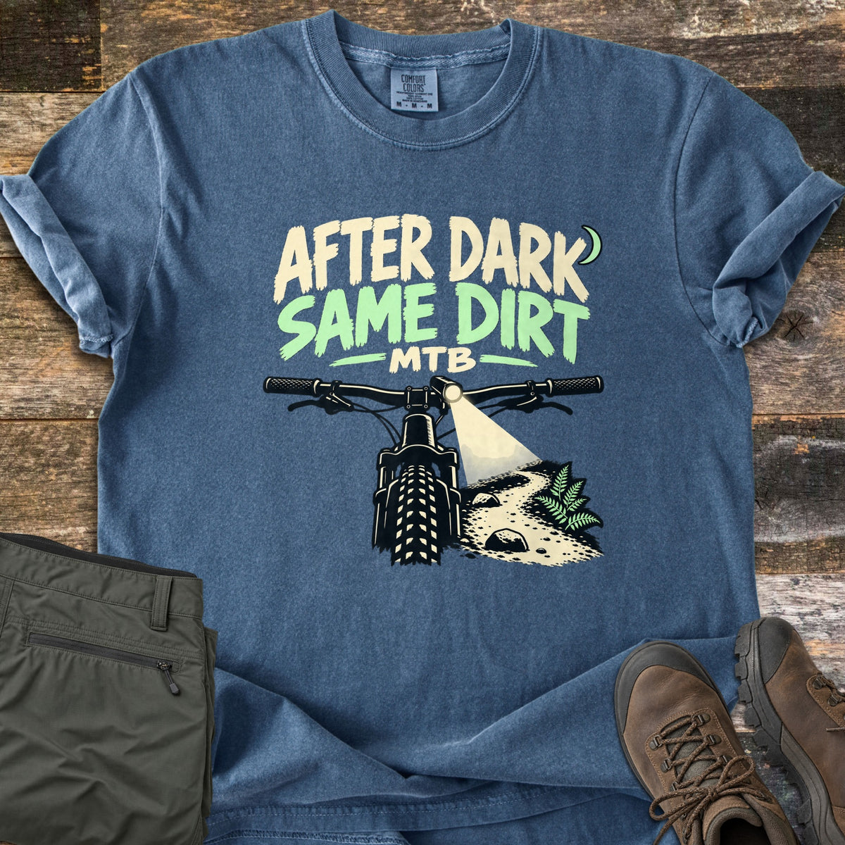 After Dark Same Dirt Comfort Colors T-shirt in Denim, laid flat. Printed words After Dark Same Dirt MTB with a crescent moon above a mountain bike's handlebars, its headlight beaming onto a dirt trail with a fern. Olive pants and brown hiking boots lie on dark wood planks.