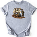 Self Parking Comfort Colors T-shirt in Sport Grey, laid flat. A cartoon rider in a helmet stands arms crossed beside a mud-caked green ATV sunk in thick mud, under the words Self Parking. The shirt lies on a white background with rolled sleeves.