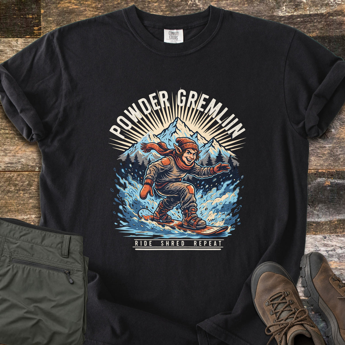 Powder Gremlin Comfort Colors T-shirt in Black, laid flat. A grinning gremlin in a knit hat and orange scarf snowboards through blue powder below snowy peaks, between the words Powder Gremlin and Ride Shred Repeat. Olive pants and brown boots sit on weathered wood.