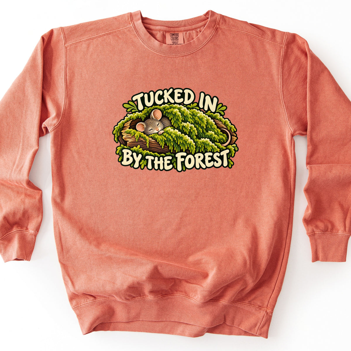 Tucked in by the Forest Comfort Colors crewneck sweatshirt in Terracotta, laid flat. A sleeping gray mouse lies on a log under a blanket of green moss, framed by the words Tucked In By the Forest in cream lettering. The sweatshirt lies on a white background.
