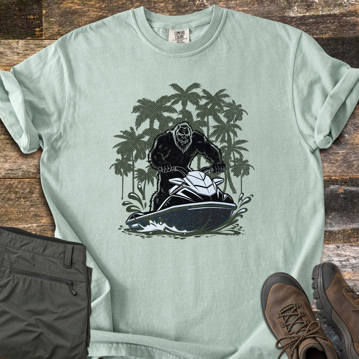 Jet Ski Squatch Comfort Colors T-shirt in Bay, laid flat. Front print shows Bigfoot riding a black and white jet ski through splashing water, with dark green palm trees behind. Sleeves cuffed, on a wood surface with olive pants and brown hiking boots.