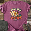 Post Ride Sniff Test Comfort Colors T-shirt in Berry, laid flat. A scruffy tan and white dog sniffs a muddy teal sneaker between the words Post-Ride and Sniff Test, with MTB and small mountains below. Folded olive pants and brown hiking boots lie beside it on wood planks.
