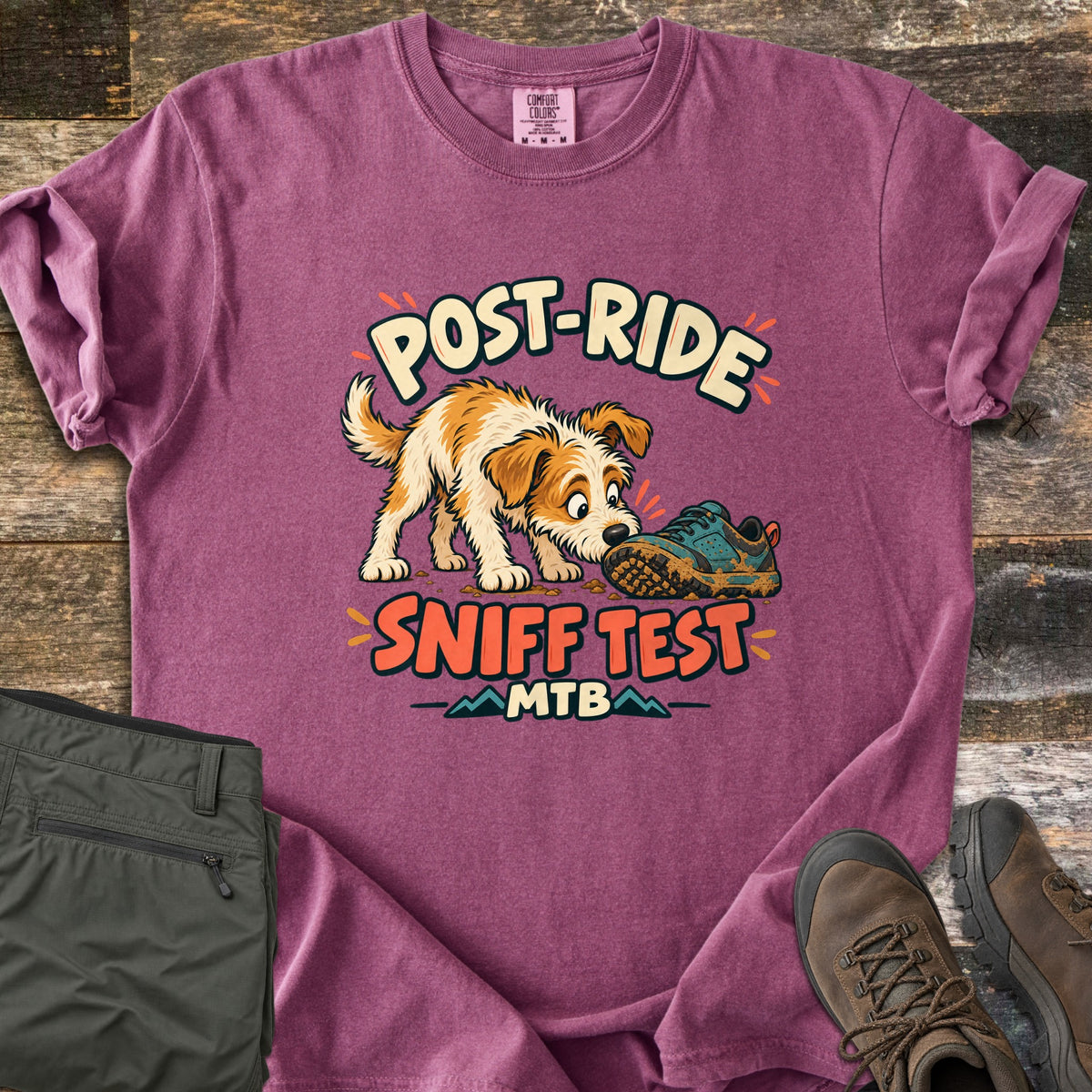 Post Ride Sniff Test Comfort Colors T-shirt in Berry, laid flat. A scruffy tan and white dog sniffs a muddy teal sneaker between the words Post-Ride and Sniff Test, with MTB and small mountains below. Folded olive pants and brown hiking boots lie beside it on wood planks.