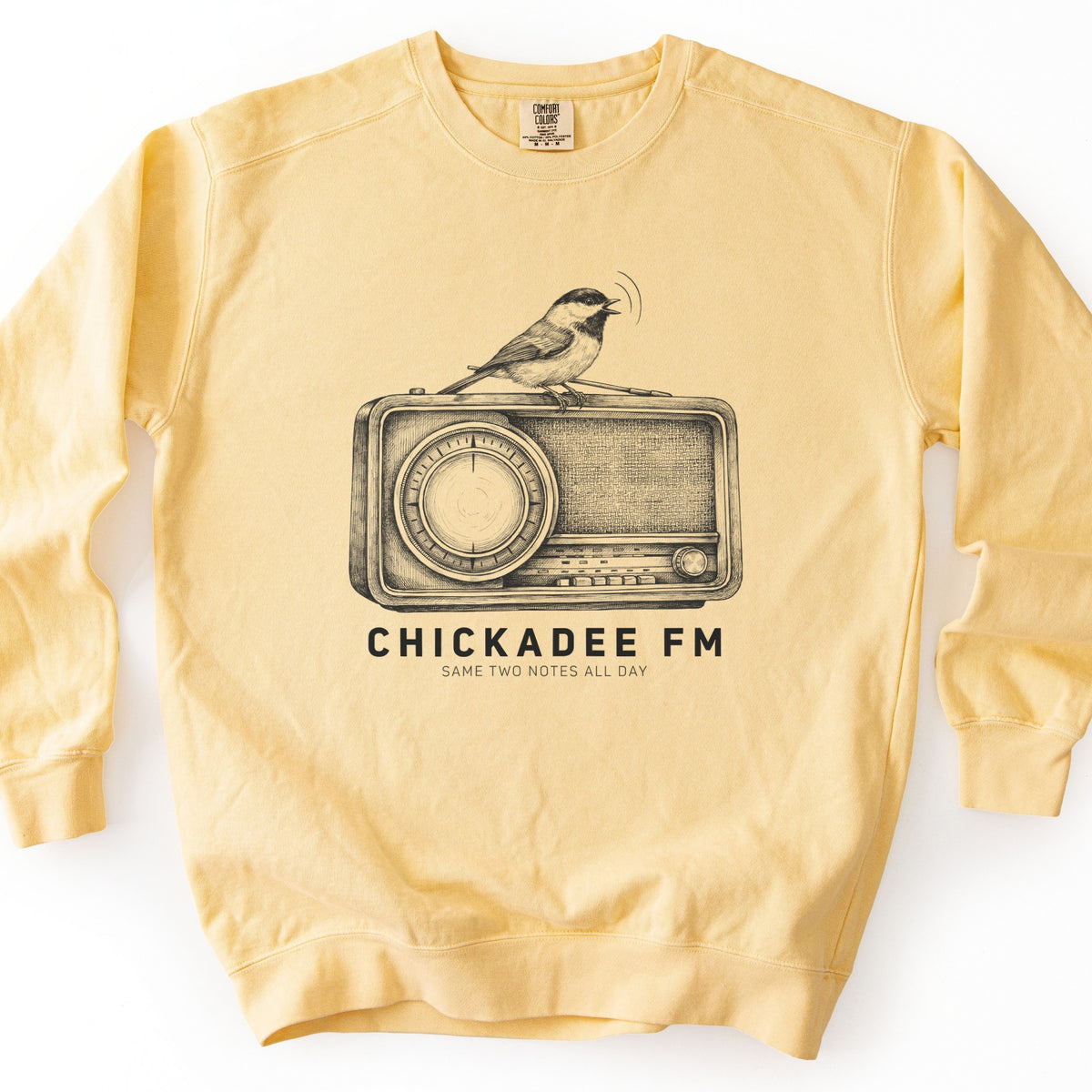 Chickadee FM Comfort Colors crewneck sweatshirt in Butter, laid flat. A sketched chickadee sings atop a vintage radio, printed in black above the words CHICKADEE FM and SAME TWO NOTES ALL DAY. It lies on a white surface with the sleeves out to the sides.