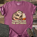 Thankful For The Leftovers Comfort Colors T-shirt in Berry, laid flat. A smiling cartoon opossum hugs a white leftover container beside a stack of containers and an orange autumn leaf, above the words Thankful For The Leftovers. Olive pants and brown boots sit on a wooden floor.
