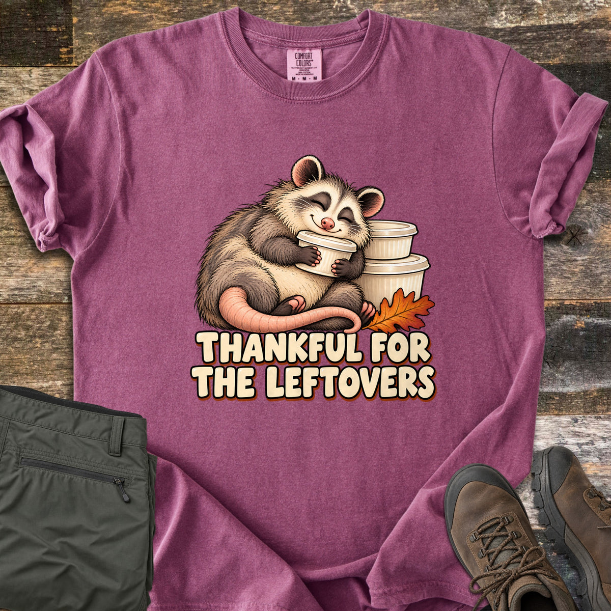 Thankful For The Leftovers Comfort Colors T-shirt in Berry, laid flat. A smiling cartoon opossum hugs a white leftover container beside a stack of containers and an orange autumn leaf, above the words Thankful For The Leftovers. Olive pants and brown boots sit on a wooden floor.