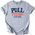Pull Please Comfort Colors T-shirt in Sport Grey, laid flat. Bold navy letters read PULL above the word PLEASE in coral script with an underline swash, and smaller navy letters below read CLAY DAYS. The sleeves are rolled and the shirt lies on a white background.