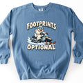 Footprints Optional Comfort Colors crewneck sweatshirt in Blue Jean, laid flat. A grinning white yeti in ski goggles rides a snowmobile through spraying snow, between snow-capped cream letters reading FOOTPRINTS above and OPTIONAL below. Shown on a white background.