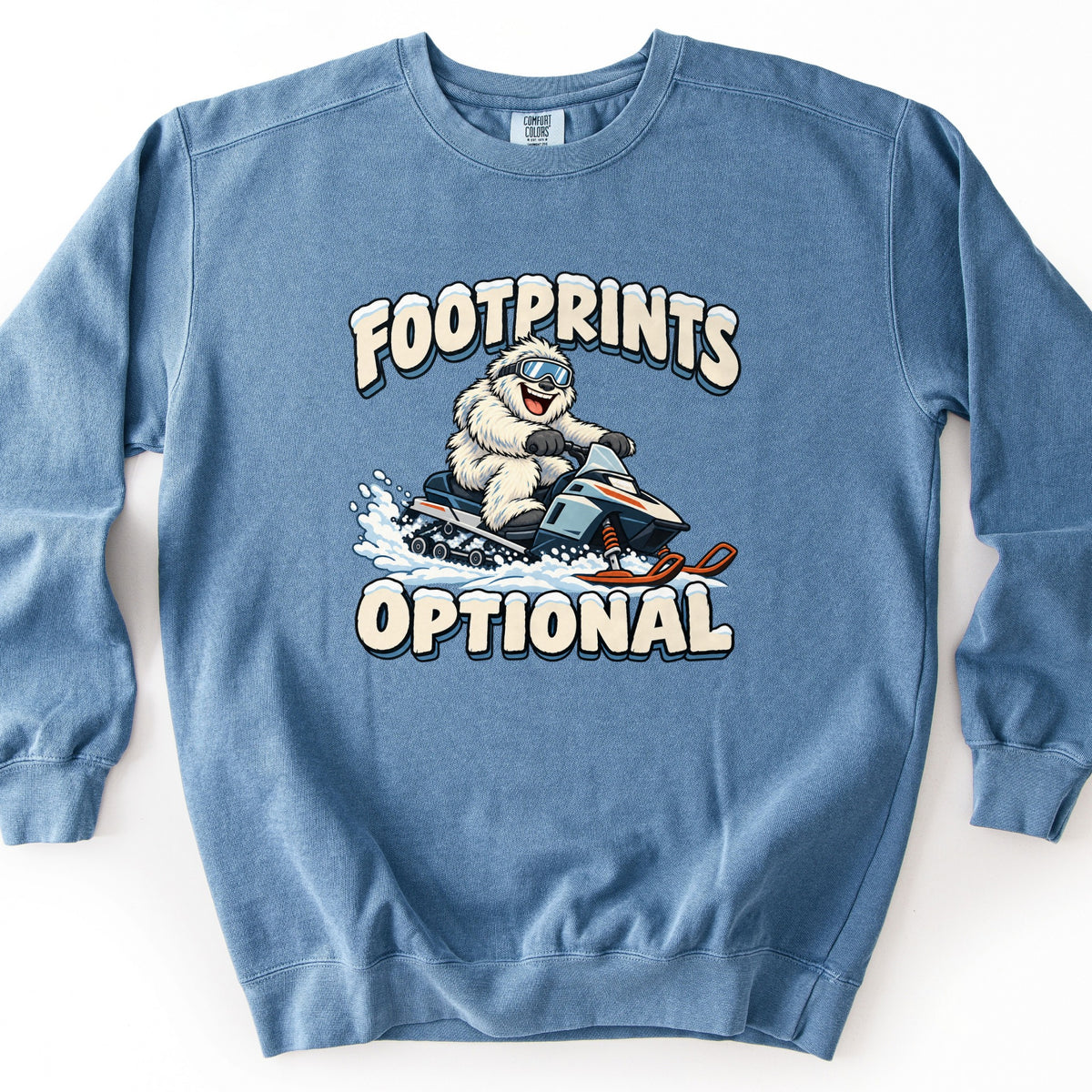 Footprints Optional Comfort Colors crewneck sweatshirt in Blue Jean, laid flat. A grinning white yeti in ski goggles rides a snowmobile through spraying snow, between snow-capped cream letters reading FOOTPRINTS above and OPTIONAL below. Shown on a white background.