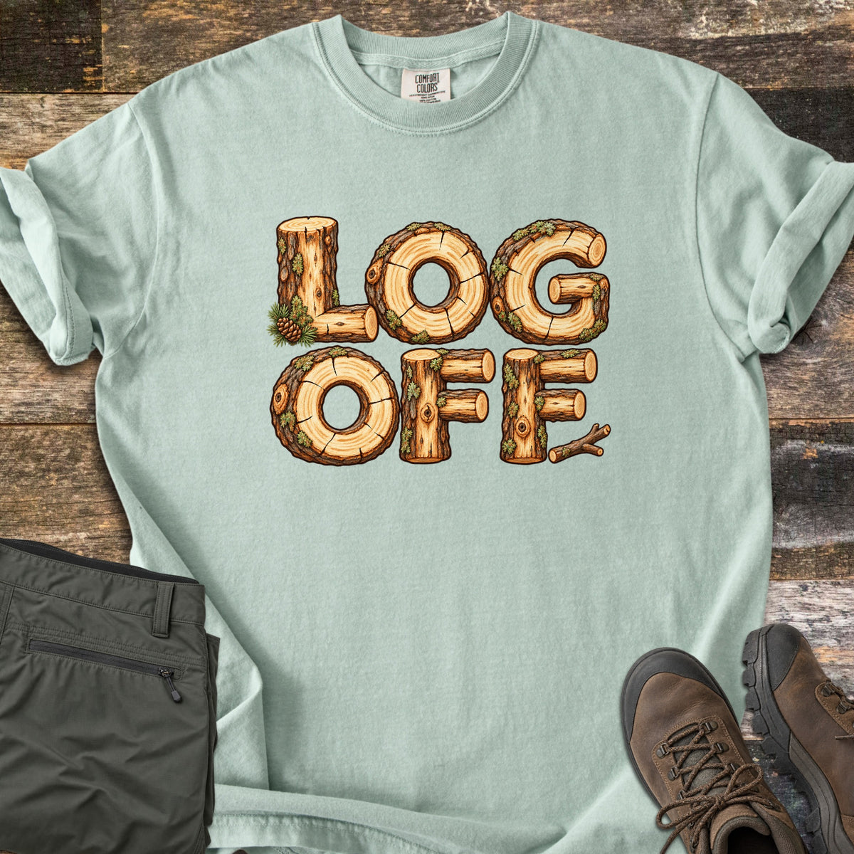 Log Off Comfort Colors T-shirt in Bay, laid flat. The words Log Off are spelled in letters made of cut wooden logs with bark, moss and a pine cone. The shirt lies on weathered wood planks beside folded olive pants and brown hiking boots.
