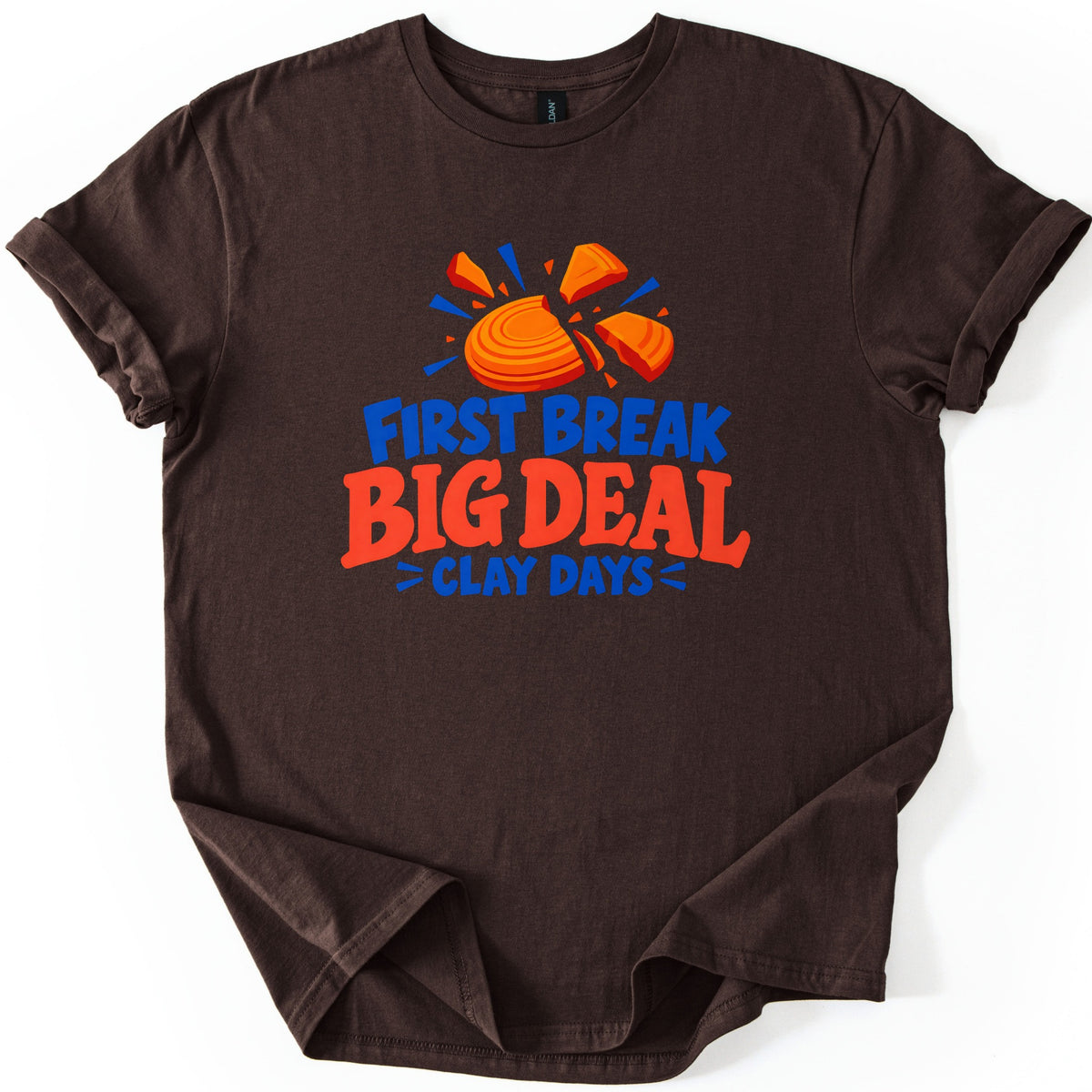 First Break Big Deal Comfort Colors T-shirt in Dark Chocolate, laid flat. An orange clay target breaking apart with blue burst lines sits above the words First Break Big Deal Clay Days, in blue and red-orange letters. The sleeves are rolled on a plain white background.