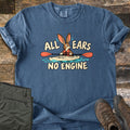 All Ears No Engine Comfort Colors T-shirt in Denim, laid flat. A cartoon rabbit in an orange life vest paddles a cream kayak with an orange-bladed paddle on blue water, between the words All Ears No Engine. Olive cargo pants and brown hiking boots sit on a wood plank floor.