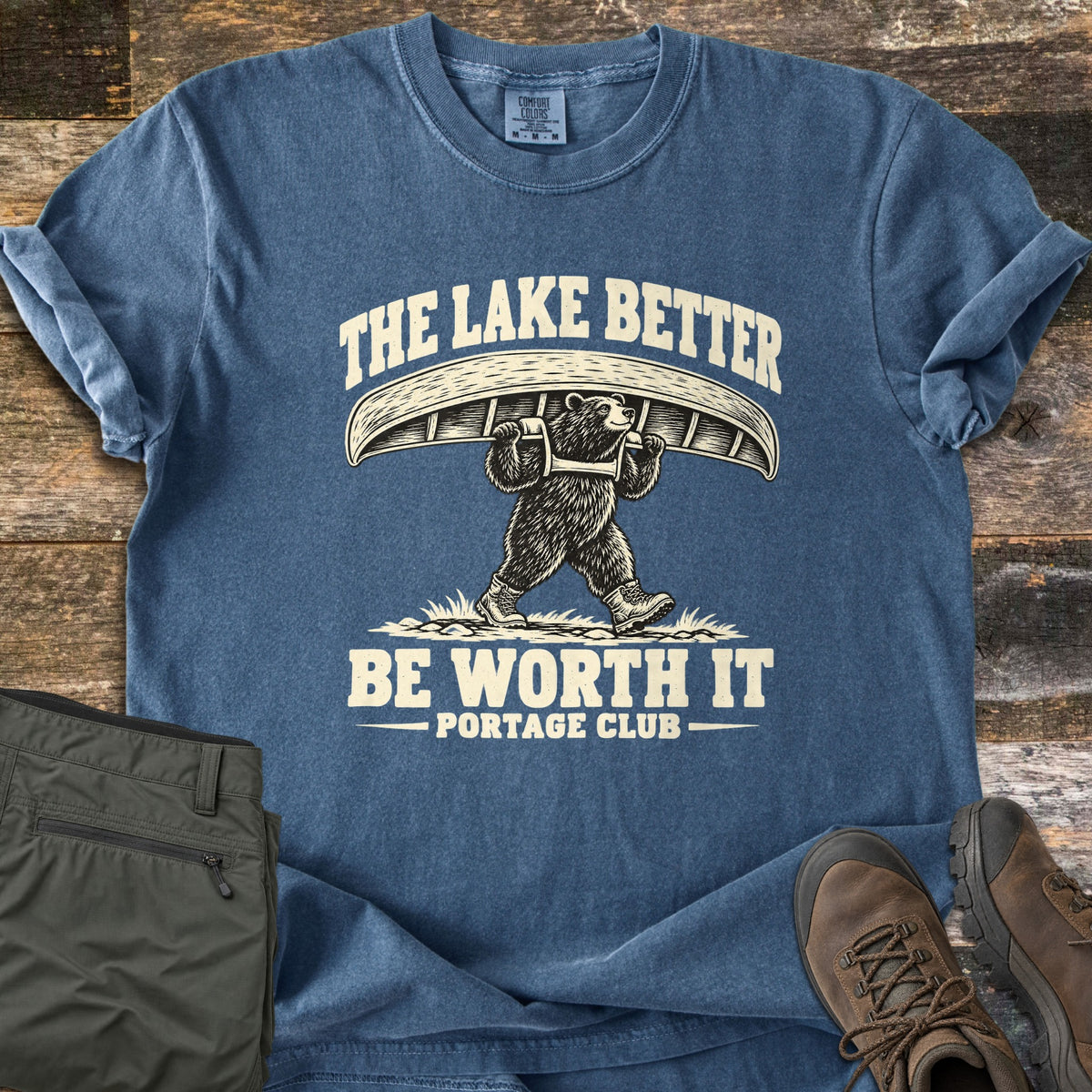 The Lake Better Be Worth It Comfort Colors T-shirt in Denim, laid flat. A bear in hiking boots carries a canoe overhead between the words The Lake Better Be Worth It, with Portage Club below, printed in cream. Shown on weathered wood with folded green pants and brown hiking boots.